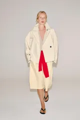 Trench corto color marfil de algodón, con cuello camisero, solapa cortaventos, mangas largas amplias con puños abotonados y cierre cruzado con un botón visible.