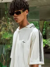 Chomba de tejido de punto cerrado color blanco, con cuello polo y mangas cortas. Presenta un bordado sutil en el frente y tres franjas negras en los hombros.