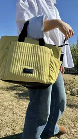 Cartera tipo tote de nylon color malva, con diseño capitoneado de líneas verticales en el frente y rombos en los laterales. Cuenta con doble asa de mano en color negro, bolsillos laterales y un parche decorativo con logo en la parte frontal.