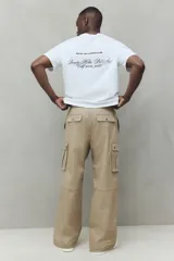 Pantalón cargo de corte holgado en color beige, confeccionado en tejido de algodón. Presenta cintura elástica con cierre de botón a presión, bolsillos laterales al bies, bolsillos de fuelle con solapa en las piernas y pliegues decorativos en las rodillas.