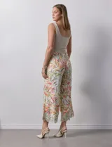 Pantalón capri de lino color crudo con estampado de hojas en tonos verdes, amarillos y naranjas. Presenta bolsillos laterales y detalle de puntilla en el ruedo.