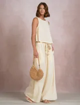Pantalón palazzo de tiro alto marca Elan, en color beige, confeccionado en mezcla de algodón, viscosa y lino. Presenta cintura delantera plana con parte trasera elastizada, bolsillos laterales funcionales y corte de pierna ancha. Incluye un cinturón trenzado al tono con terminación en borlas y flecos.