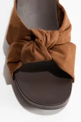Sandalias estilo zueco color marrón, con tiras de gamuza anudadas en la parte delantera y detalles de tachas metálicas doradas en los laterales. Cuenta con suela de madera oscura de tacón cuadrado bajo.
