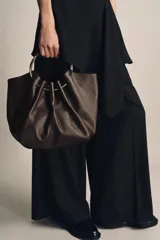 Bolso tipo saca de piel color marrón oscuro, con cuerpo fruncido y doble asa de mano rígida metálica. Incluye asa extraíble para llevar al hombro o cruzado y bolsillo interior extraíble.