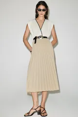 Vestido midi bicolor con parte superior blanca de escote en V cruzado con ribete negro y falda plisada en color tostado. Cuenta con mangas sisa, cintura elástica y un cinturón negro tipo reptil con hebilla ovalada.