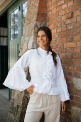 Blusa de lino blanca con cuello alto, volados frontales y mangas largas amplias con terminaciones en volado.