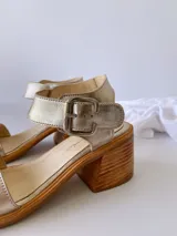 Sandalias de cuero color dorado, con tira ancha sobre los dedos, pulsera al tobillo con hebilla metálica y taco de madera.