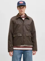 Campera de estilo worker color marrón, con cuello camisero de pana, cierre frontal con cremallera y bolsillos frontales con solapa y broches a presión. Presenta un acabado recubierto que le otorga una textura sutilmente brillante y resistente.