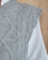 Chaleco de punto gris con diseño de trenzas y rombos en relieve, cuello redondo y terminaciones acanaladas.