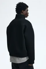 Buzo negro de tejido pesado con efecto neopreno, cuello elevado y medio cierre frontal plateado. Presenta hombros caídos, mangas largas y puños y bajo acanalados.