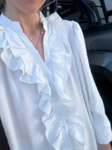 Vestido largo color crudo, estilo camisero, con cuello en V y volados verticales en el frente. Tiene mangas largas abullonadas y corte amplio.