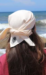 Conjunto compuesto por un pareo corto de color beige con cierre cruzado y detalle de bordado, y una bandana a juego. El pareo presenta un diseño envolvente con lazo lateral.