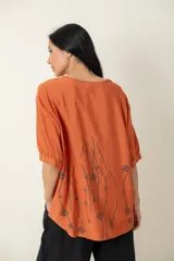 Blusa color ladrillo con estampado de flores y líneas negras, de corte holgado, cuello redondo y mangas cortas con puños elásticos.