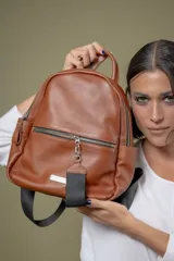 Mochila de cuero vacuno color suela, con diseño compacto y funcional. Cuenta con un compartimento principal con cierre, un bolsillo frontal con cierre y tirador de cinta negra, y correas ajustables de cinta. El interior está forrado en tela de algodón.