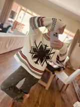 Sweater tejido artesanalmente en lana color crudo, con diseño de copo de nieve negro en el frente y la espalda. Presenta detalles de punto en color naranja y negro en cuello, puños y ruedo. Mangas largas con diseño de punto calado en los hombros.