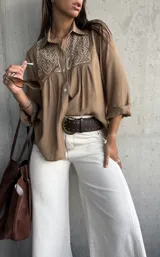 Camisa de color camel con cuello clásico, manga larga y detalle de bordado geométrico en color blanco en la parte frontal superior. Presenta cierre frontal con botones y un corte holgado.