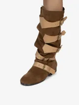Bota de montar de caña alta y diseño holgado, confeccionada en cuero con acabado de gamuza color marrón. Presenta múltiples correas de cuero liso en color beige con hebillas metálicas doradas a lo largo de la caña y el empeine. Posee un taco bajo de aproximadamente 2,5 cm.