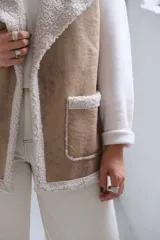 Chaleco de gamuza color beige con interior y detalles de corderito color crudo. Presenta solapas amplias, bolsillos frontales aplicados y diseño sin mangas.