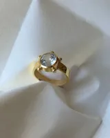 Anillo de banda ancha con baño de oro, con una piedra facetada central transparente y redonda, rodeada por pequeños detalles esféricos.