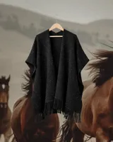 Ruana tejida en telar con lana merino, color negro con sutiles destellos brillantes. Presenta un diseño abierto con flecos en el ruedo.
