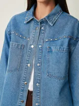 Camisa de denim celeste oversize con bolsillos frontales, cierre de botones metálicos y detalles de tachas en el cuello y la parte delantera.