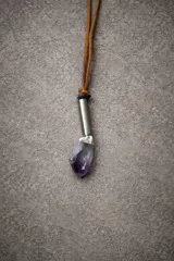 Collar con tiento de cuero marrón y dije colgante que consiste en un cristal de amatista en bruto engarzado en una pieza metálica cilíndrica plateada.