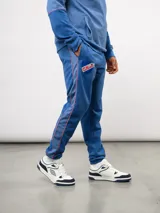 Pantalón deportivo de piqué color azul marino, con franjas laterales en contraste rojo y blanco. Presenta el escudo del Club Nacional de Football bordado en la pierna derecha y una etiqueta conmemorativa en la izquierda.