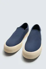 Zapatilla tipo slip-on de lona color taupe, con elástico lateral para facilitar el calce y suela de goma gruesa color crema.