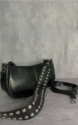 Cartera tipo baguette de cuero sintético negro con acabado brillante, cierre superior y correa ancha decorada con tachas metálicas plateadas.