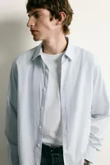 Camisa de corte regular fit confeccionada en tejido de algodón con diseño de rayas verticales finas. Presenta cuello italiano, manga larga con puños abotonados y cierre frontal mediante botonadura.
