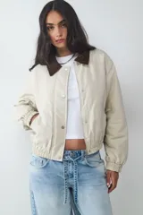 Campera acolchada corta color beige, con cuello de pana en contraste color marrón. Cuenta con cierre frontal de cremallera y botones a presión, bolsillos laterales, y puños y bajo elásticos.