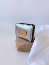 Billetera pequeña de cuero color plateado con acabado metalizado, cierre perimetral metálico y parche de cuero en tono natural con el logo de la marca grabado.