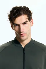 Campera bomber deportiva de tejido funcional color verde oscuro, con cuello elevado corto, cierre bidireccional y mangas raglán. Posee bolsillos delanteros con cierre y costuras selladas.