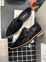 Zapato acordonado de cuero charol negro con detalle de trenzado en la puntera y suela color beige.