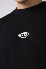 Remera de corte oversize confeccionada en algodón pesado, color negro. Presenta un bordado minimalista en el centro del pecho con forma de ojo y un parche estampado en la espalda que ilustra una mano abriendo una persiana.