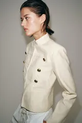 Chaqueta estilo militar ZW Collection confeccionada en hilatura con mezcla de algodón, color crudo, con cuello subido, manga larga, falsos bolsillos de vivo y cierre frontal con ganchos metálicos y botones decorativos dorados.