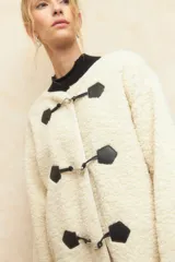 Abrigo gamulán de corderito color crudo, con alamares de cuero sintético negro y cierres metálicos.