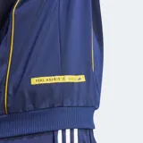 Campera deportiva Adidas de hombre, color azul marino, inspirada en la equipación del Real Madrid temporada 99/00. Presenta cuello alto, cierre frontal, tres franjas blancas en las mangas y ribetes amarillos en los costados. Lleva el escudo del Real Madrid y el logo de Adidas bordados en el pecho.