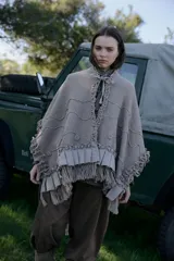 Ruana de tejido de punto con textura, color beige, con cuello alto con volados y lazo de ajuste frontal. Presenta detalles de volados aplicados en lino en el ruedo y mangas, y terminación con flecos artesanales.