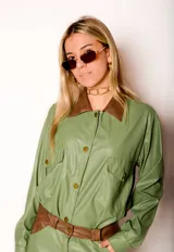 Camisa verde de cuero sintético con cuello marrón, cierre frontal con botones, dos bolsillos con solapa en el pecho y cinturón marrón.