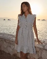 Vestido corto de lino color gris claro, sin mangas, con volados en los hombros, cuello mao y cierre frontal con botones de nácar. Presenta una falda con vuelo y detalle de volado en el ruedo.