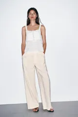 Pantalón de tiro alto en color beige, con diseño de pierna ancha y pinzas frontales. Cuenta con bolsillos laterales, cierre frontal con cremallera y gancho metálico, y un lazo ajustable en la cintura del mismo tejido.
