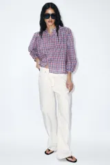 Blusa de manga larga con diseño a cuadros en tonos violeta, rosa y blanco. Presenta cuello redondo, cierre frontal con botones y mangas con frunces en los hombros.