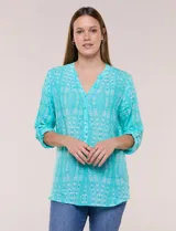 Blusa turquesa de silueta suelta confeccionada en tejido liviano, con escote en V, pequeña cartera frontal con botones decorativos y mangas 3/4 con presilla para arremangar. Presenta patrón estampado geométrico tipo tie-dye y ruedo curvo.