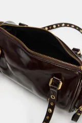 Bolso estilo bowling de cuero sintético acharolado color marrón, con diseño cilíndrico, bolsillos laterales con cierre y doble asa decorada con tachas metálicas doradas.