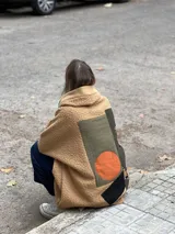 Campera tipo hoodie confeccionada en tejido de corderito color camel, con diseño de collage en la espalda que combina parches de tela en tonos verde oliva, naranja y estampados étnicos. Presenta un corte holgado y capucha integrada.