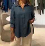 Camisa de jean de corte oversize, con cuello clásico, cierre frontal de botones y mangas largas. Presenta un diseño con bolsillos laterales integrados y ruedo redondeado.
