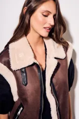 Chaleco oversize estilo gamulán en color marrón, confeccionado en cuero vegano con forro y solapas de corderito sintético en contraste. Posee cierre frontal metálico, bolsillos con cremallera y detalles de correas con hebillas en el cuello.