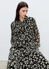 Blusa de gasa con estampado floral de margaritas sobre fondo negro, diseño recto semitransparente con cuello pico y detalle de lazo frontal. Cuenta con mangas largas y forro interior.
