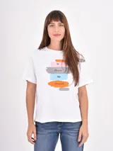 Remera blanca de algodón con cuello redondo y mangas cortas. Presenta un estampado frontal con pinceladas de colores y la frase 'in love we trust'.